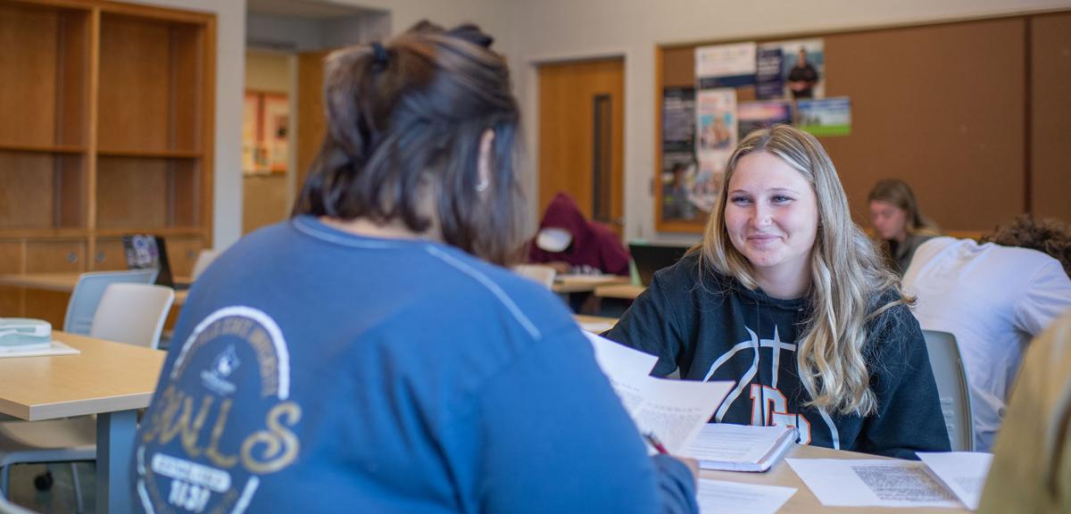 Two students work on coursework together while seated at a table in a classroom.