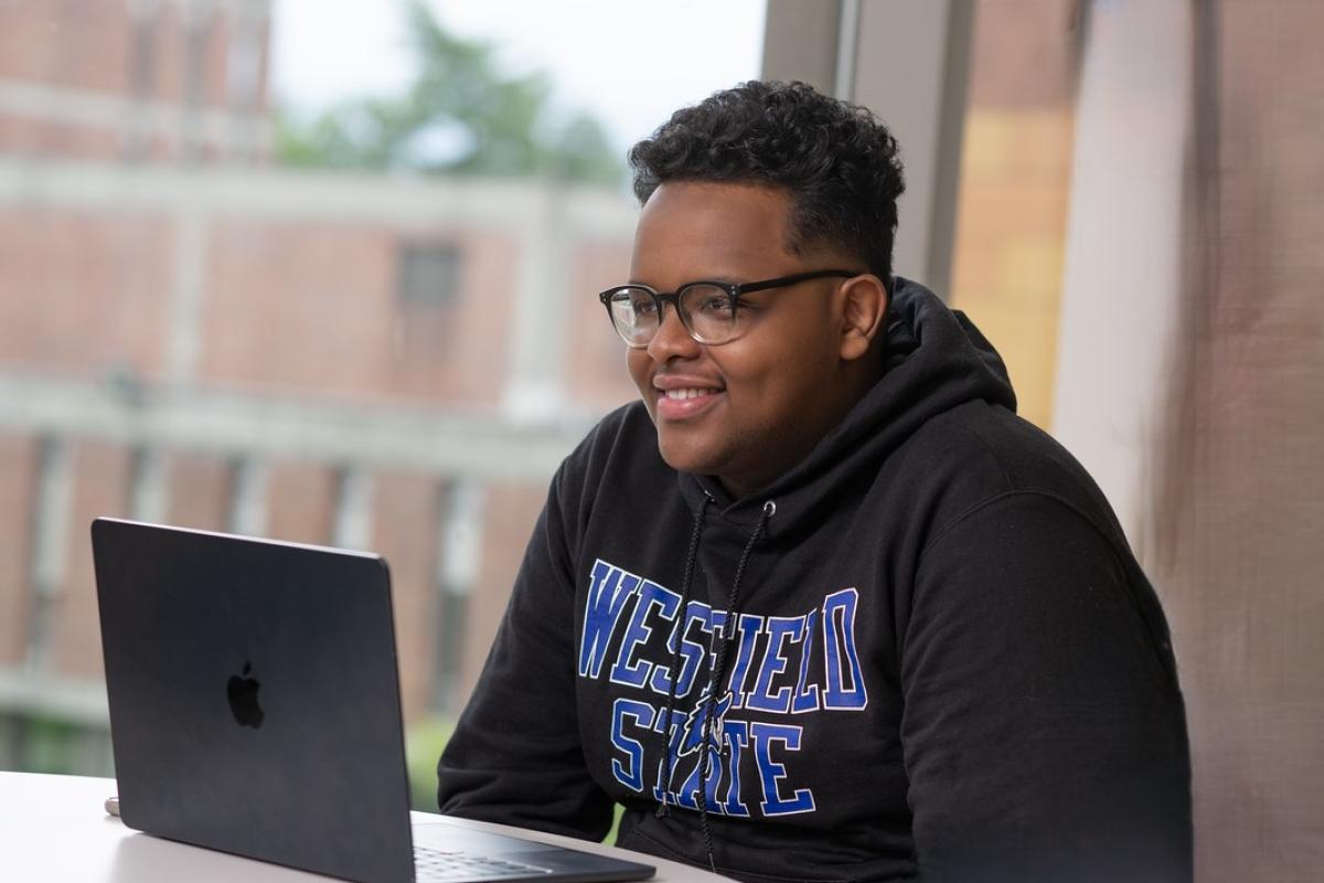 Student sitting at table with laptop smiling wearing a WSU sweatshirt.