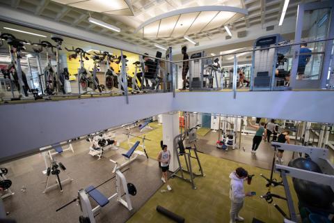 A shot of the Ely Fitness Center, shot from halfway on the staircase leading up and down. White cardio machines are stacked in a row on the upstairs portion of the room with white benches down below for weight lifting.