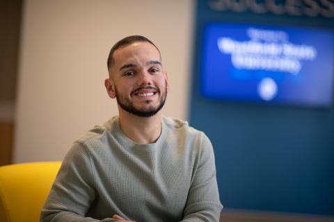 Nate Ferreira, sitting on a yellow chair with a blue and white background behind him that is out of focus.