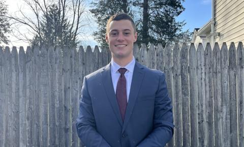 Bennett Kelly, a criminal justice major, is dressed in a blue shirt and smiles at the camera. He is outside, and a brown, wooden fence is behind him.