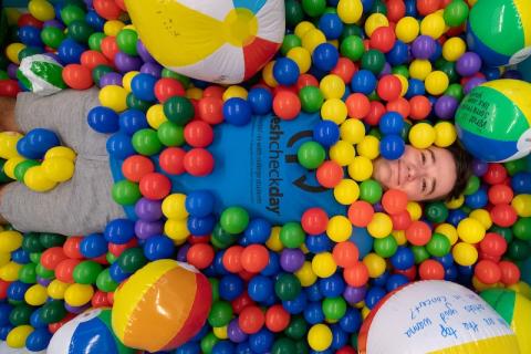 A male student from the 2023 Fresh Check Day. He is wearing a dark blue shirt with black lettering that says, "Fresh Check Day", and is laying in a colorful ball pit while smiling at the camera.