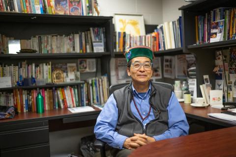 Professor Max Saito of the Communication Department. He sits in his office, which is walled with several shelves of books. His hands are clasped over his torso, and he smiles at the camera in a vest and green hat.