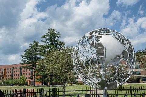 A photo of the campus globe during the summer season. The sky above is blue with clouds, and University Hall can be seen behind the globe itself.