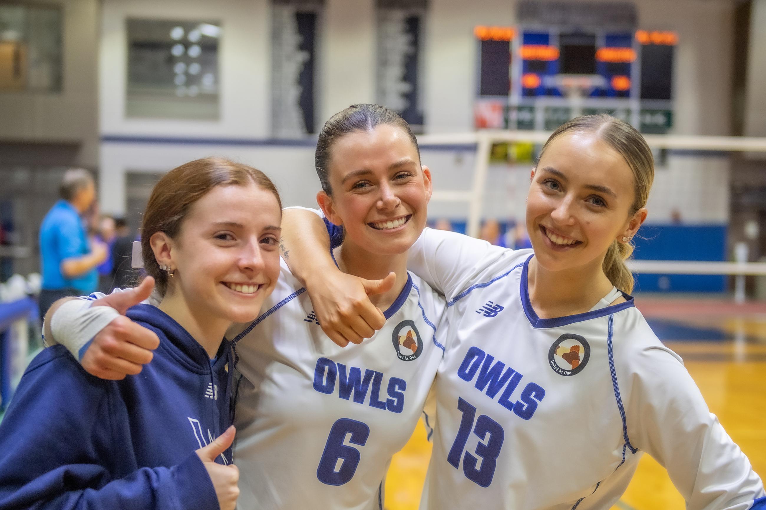 3 Westfield State University volleyball players in uniform, smile at the camera with their arms around each others shoulders