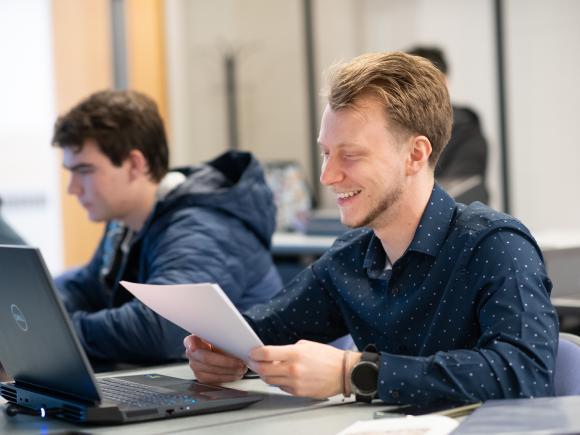 Computer science student smiling in front of lap top wearing long sleeve navy shirt.