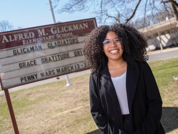 Reach to Teach alumna Lisha standing outside school smiling wearing a black coat, white shirt, and glasses.