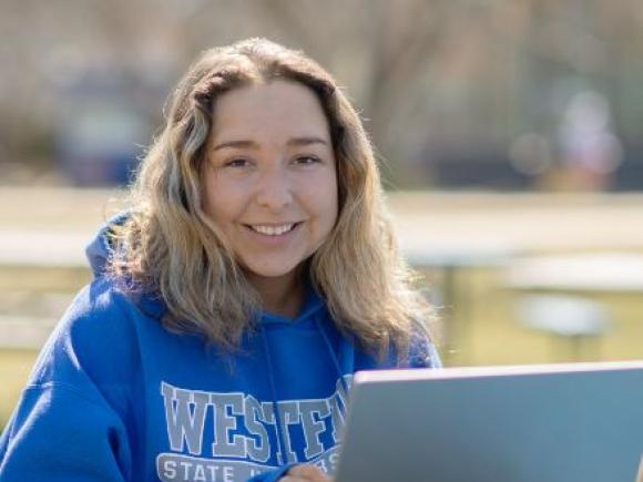 A student looks up from their laptop and smiles while sitting at a table outside.