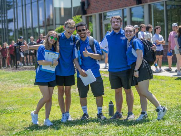 New Student Orientation. Five student leaders in blue polo shirts pose on the campus green.