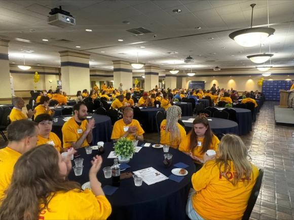 The Western MA Caring Force Rally, held on September 17 at Westfield State University’s Scanlon Hall. People in yellow shirts are seated at banquet tables and eating while discussing legislative priorities important to the rally.