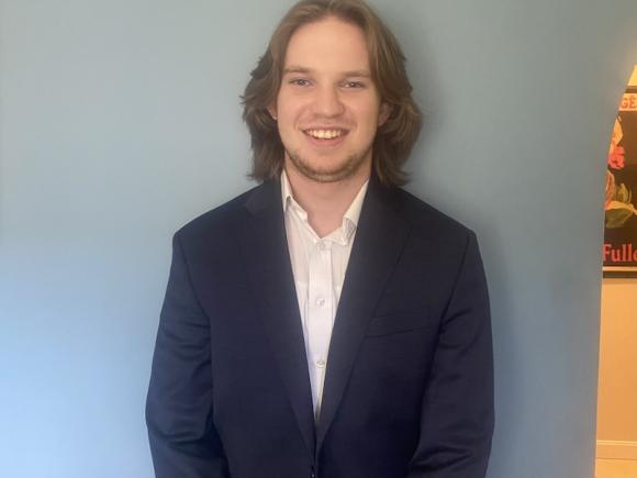 Austin Lourake, Class of 2025. He is in a blue coat jacket and white shirt and is posing for a headshot in front of a light blue wall.