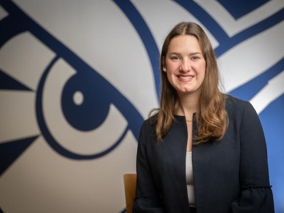 Elin-Maree Krupa, an international transfer student from Germany. She poses in front of a blue and white Owl mural and wears a dark blue coat jacket.