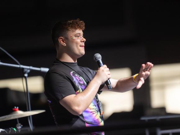 Student speaks into a microphone onstage during Westfield State’s Spring Weekend, gesturing to the audience while wearing colorful face gems.