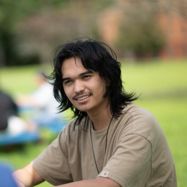 Communication student sitting out on the campus green wearing a brown shirt smiling.