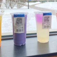 Four boba tea, orange, purple, clear, and red colored, rest on a black windowsill which overlooks the campus green. There's fresh snow on the ground and shrubbery in the background next to the stone walkway.