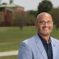 A landscape portrait of Junior Delgado, Director of the Career Center. He's wearing a navy shiny and light blue suit jacket and is standing in front of the Campus Green, which is blurred behind him.